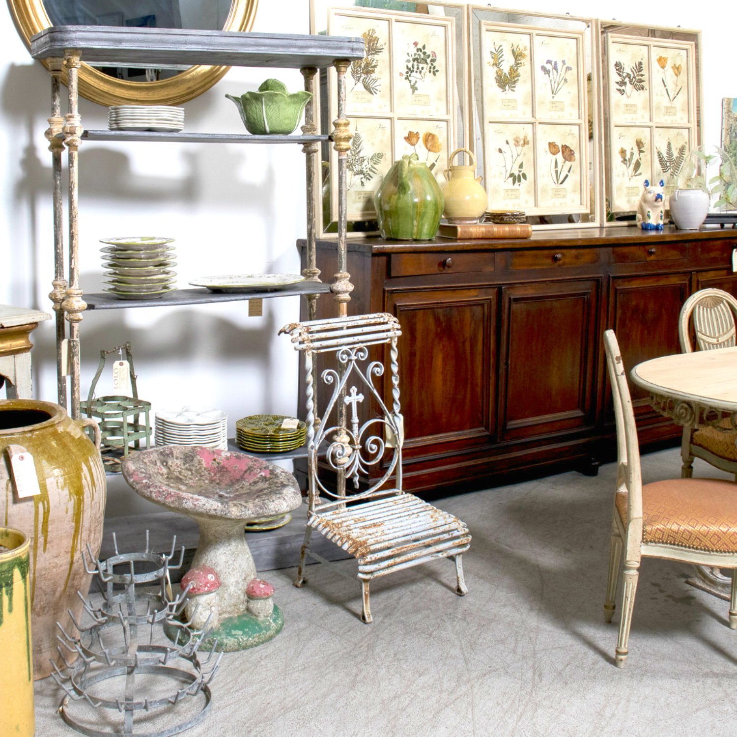 A collection of antique French oyster plates and platters displayed on a rustic étagère, surrounded by vintage garden pottery, botanical prints, and Provençal furniture at Lolo French Antiques.