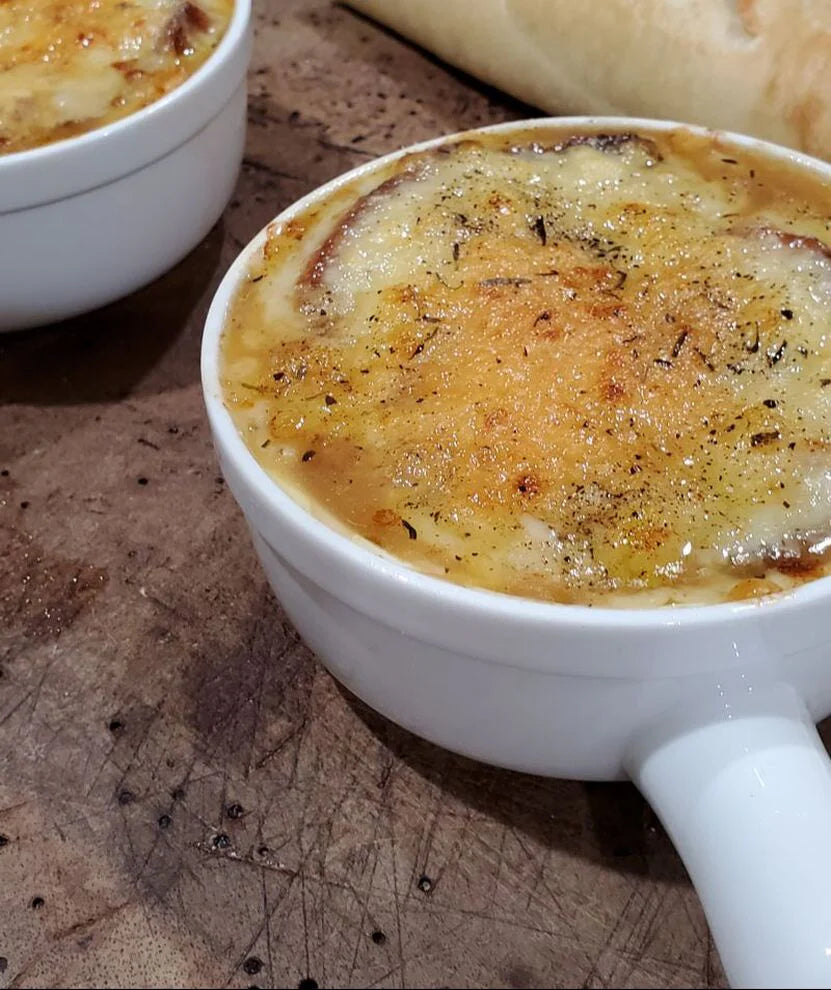 Two white crocks of French onion soup, topped with golden-brown melted cheese and sprinkled with pepper, resting on a rustic wooden surface.